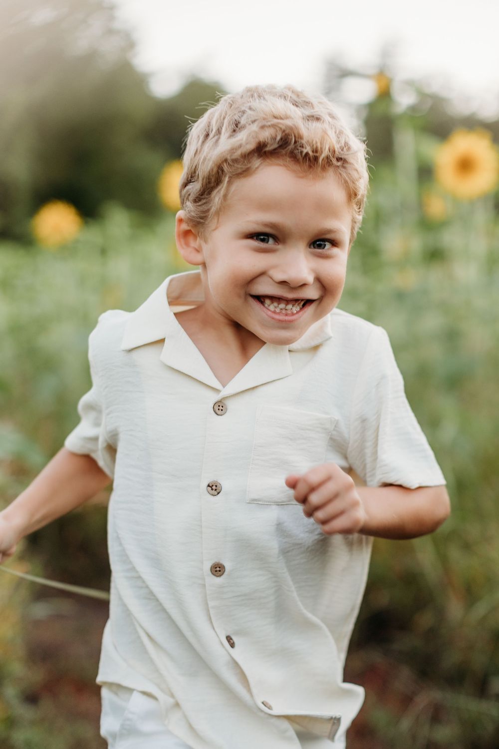 Child in white button-up shirt playing and smiling in a sunflower field.