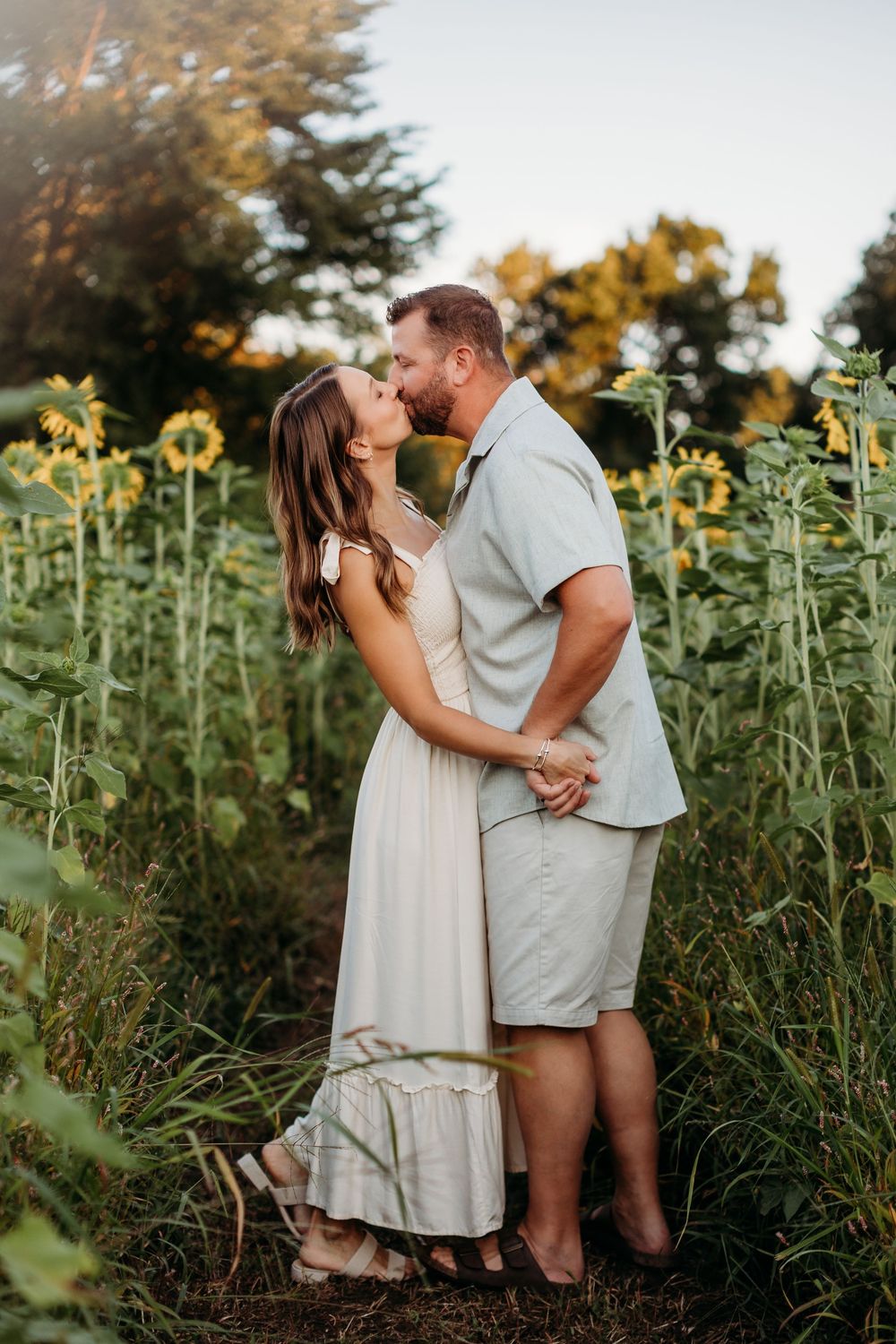 Couple in light colored outfits sharing tender moments among tall sunflowers.