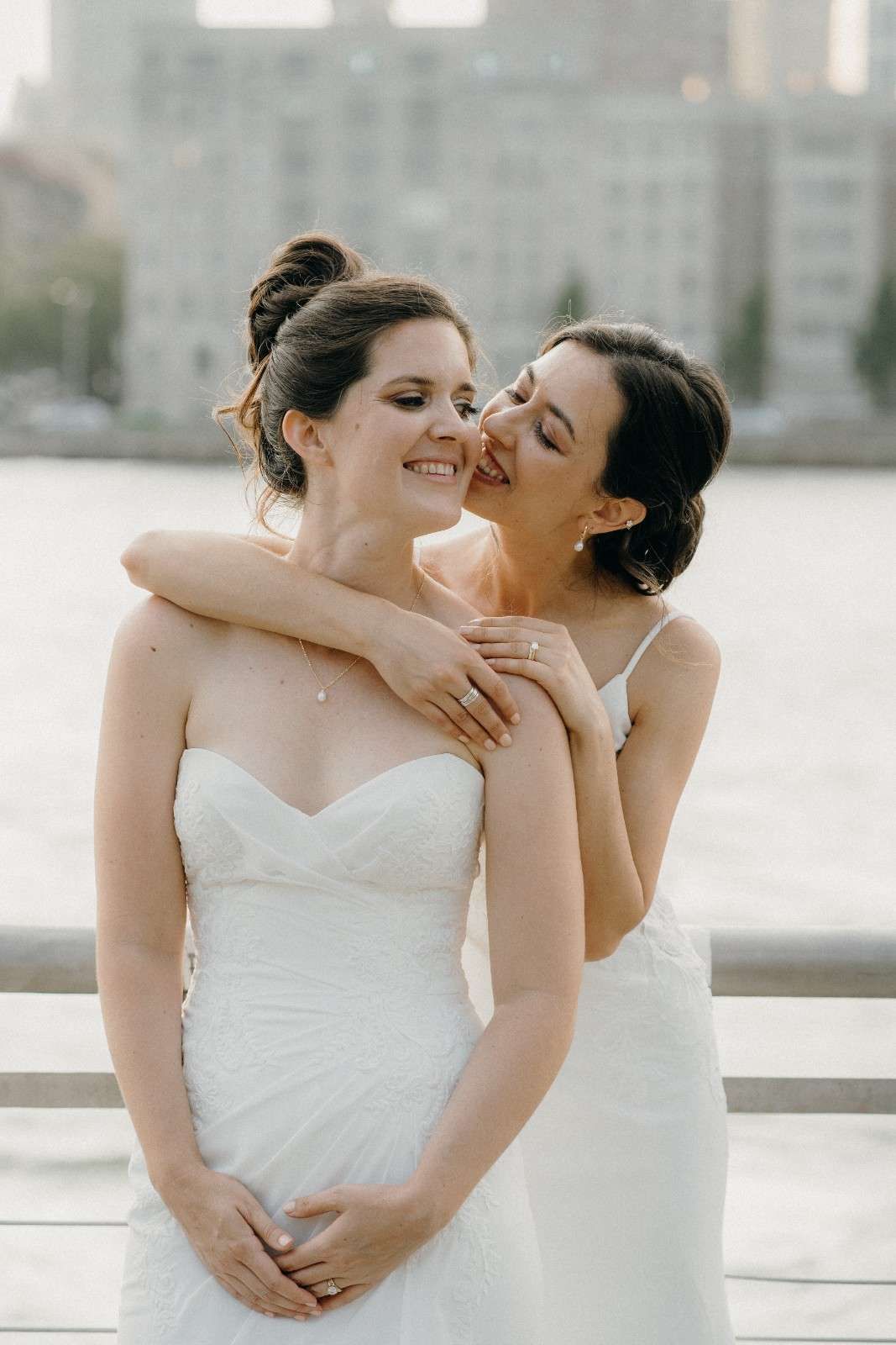 Two brides in white wedding dresses share a joyful embrace during their wedding photoshoot at the Sanctuary Roosevelt Island NYC