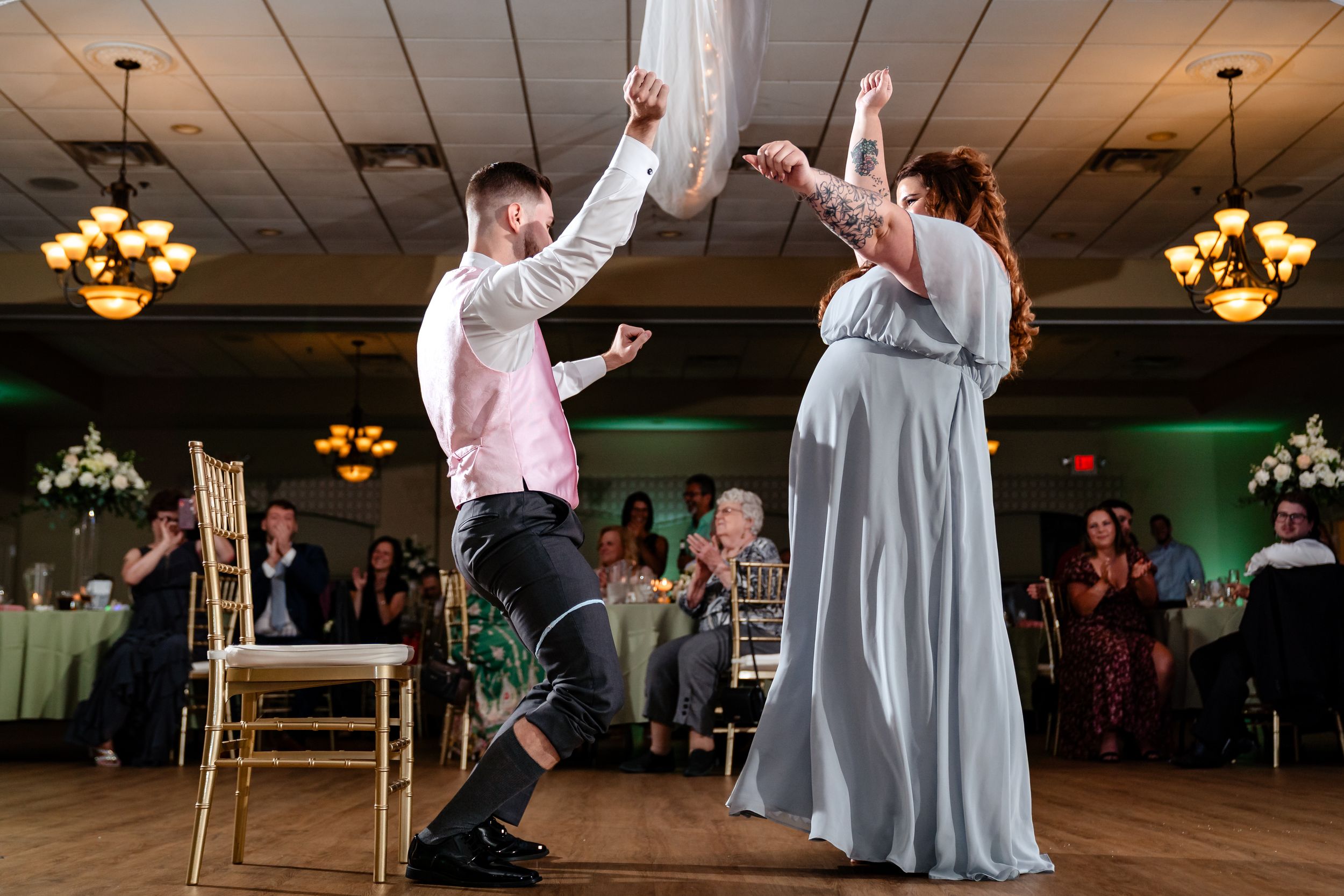 Wedding guests dancing and celebrating on a reception dance floor with chandeliers overhead.