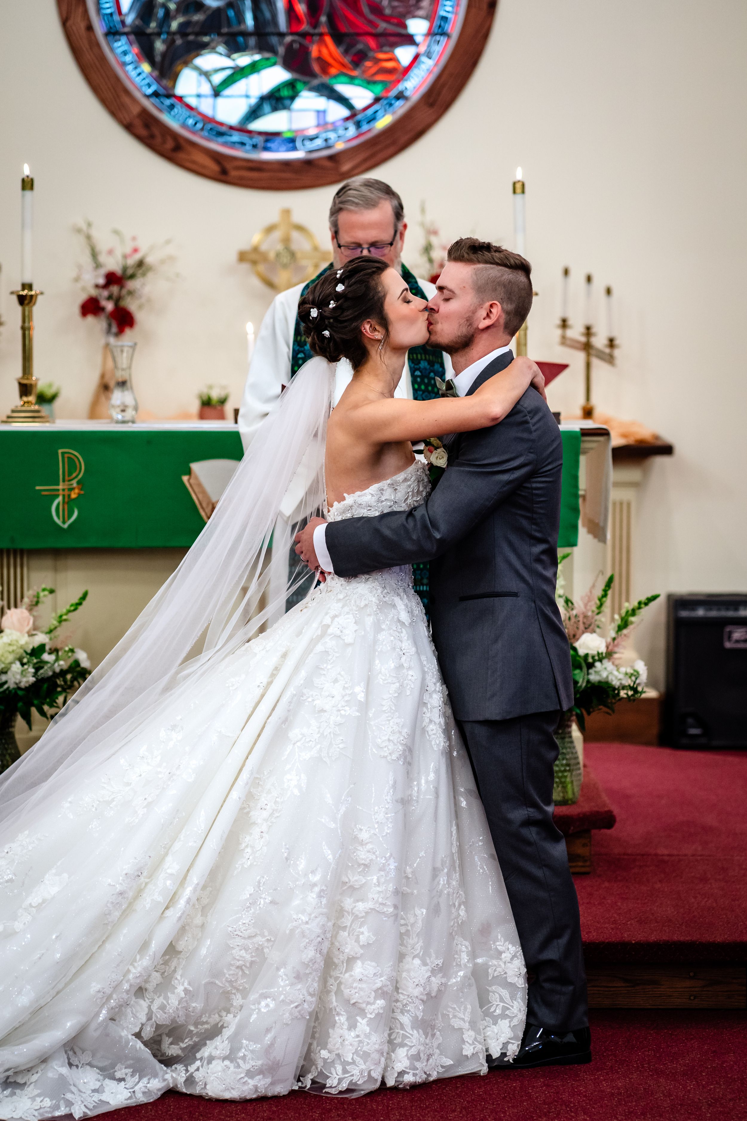 Romantic first kiss as newlyweds in church with stained glass windows and red carpet.