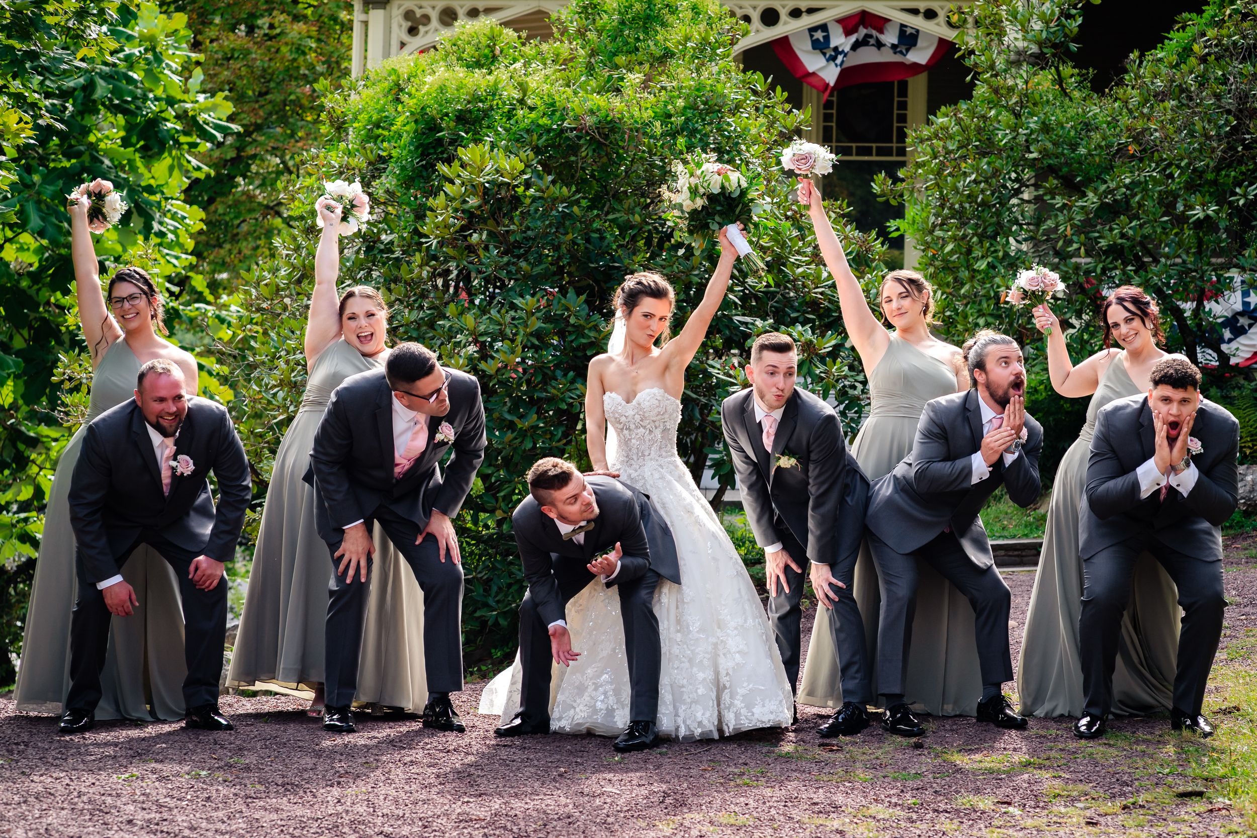 Wedding party poses playfully on grassy lawn during outdoor celebration.