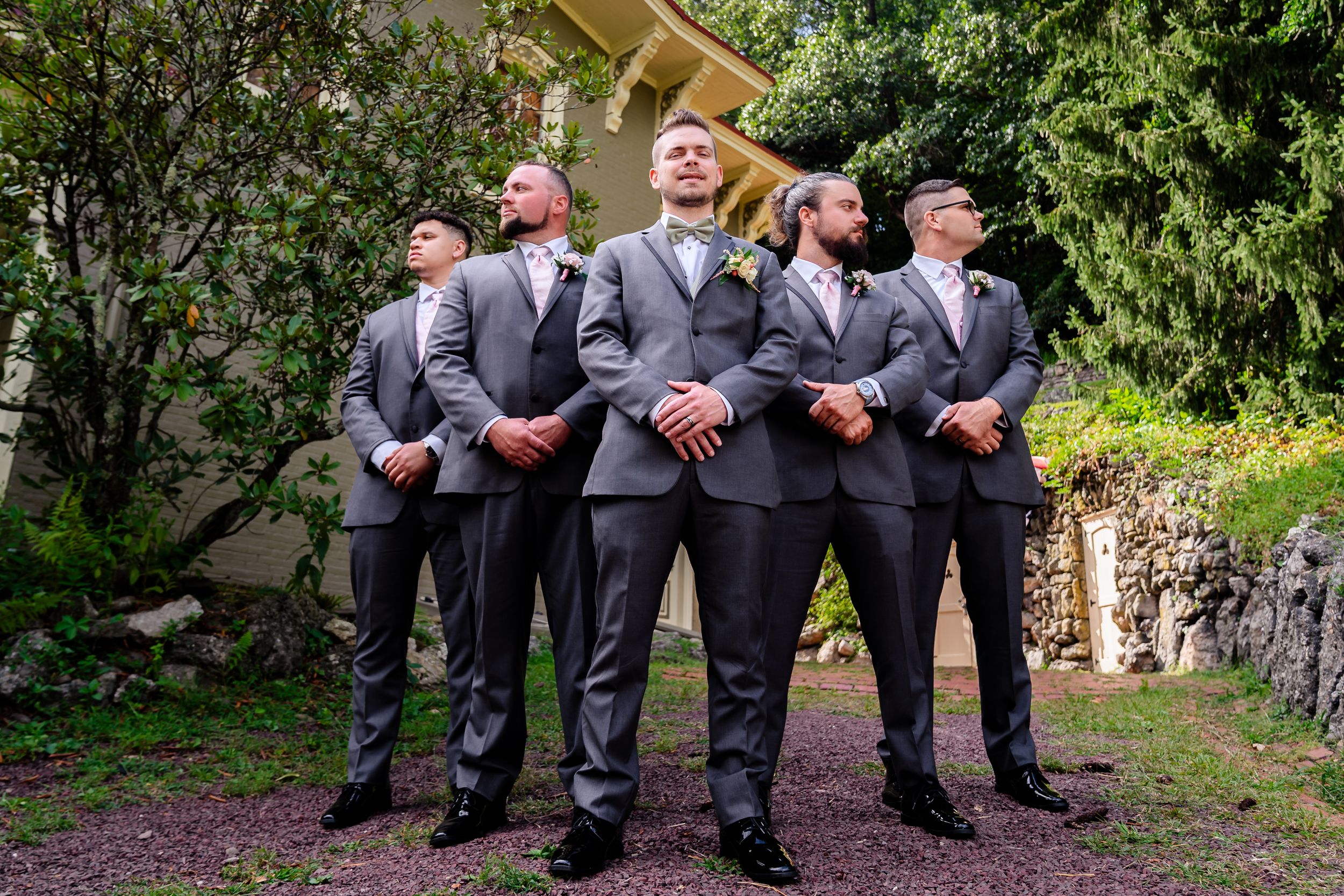 Groomsmen in dark suits pose casually outdoors for wedding photos.