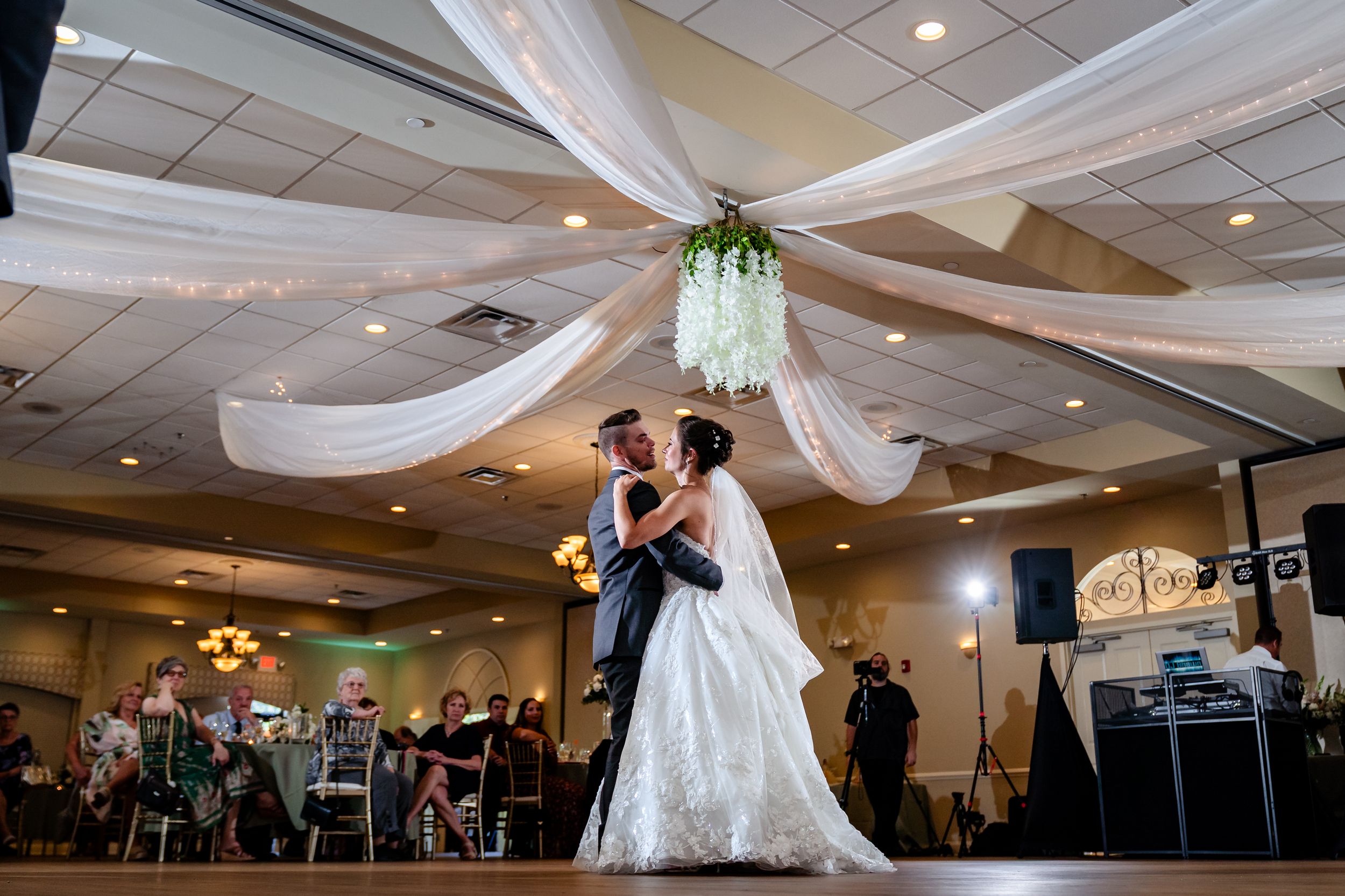 Wedding couple shares an intimate dance under elegant white fabric draping and chandeliers in a reception hall.