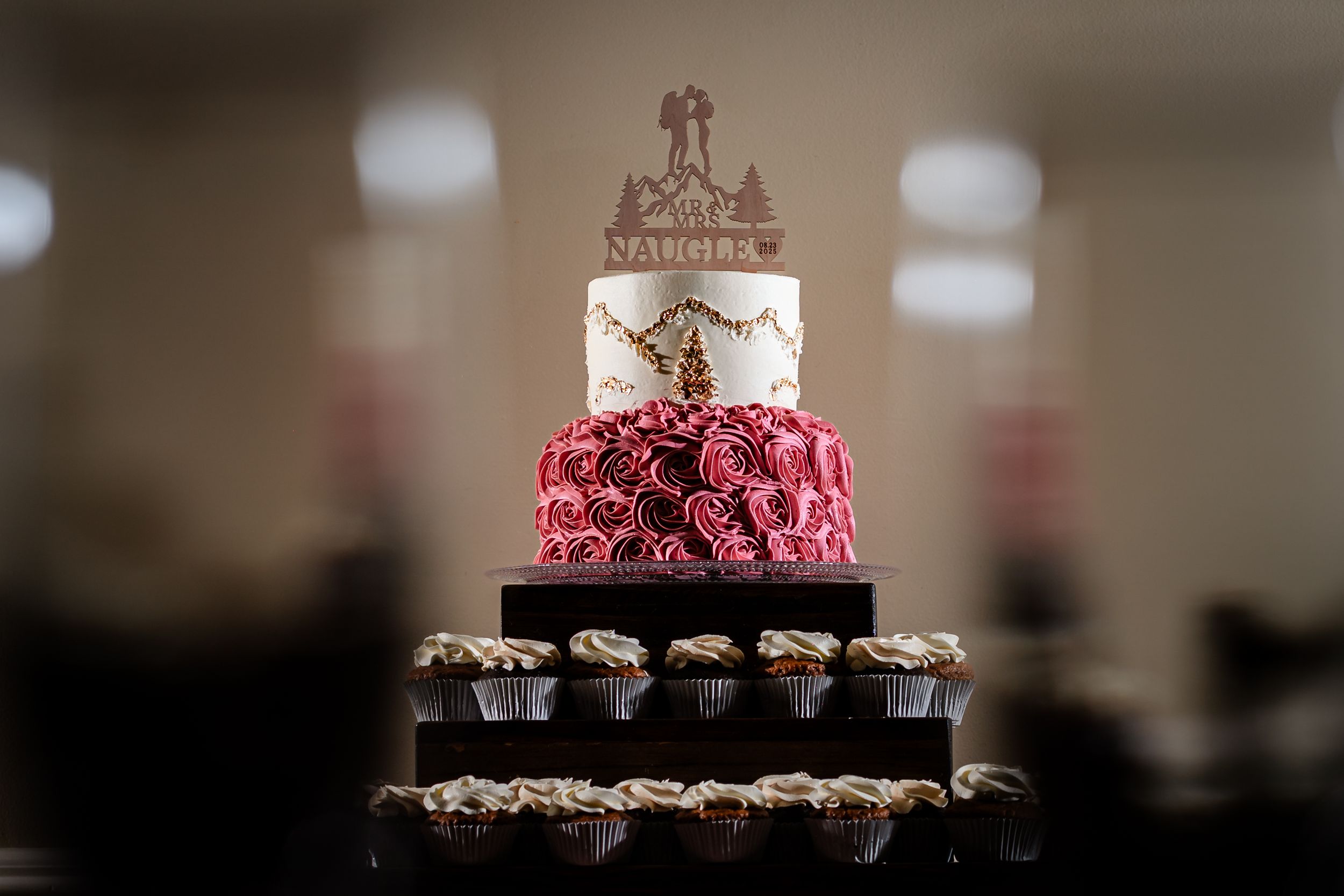 Elegant three-tier wedding cake with red rose details and cupcakes displayed on a dessert table.