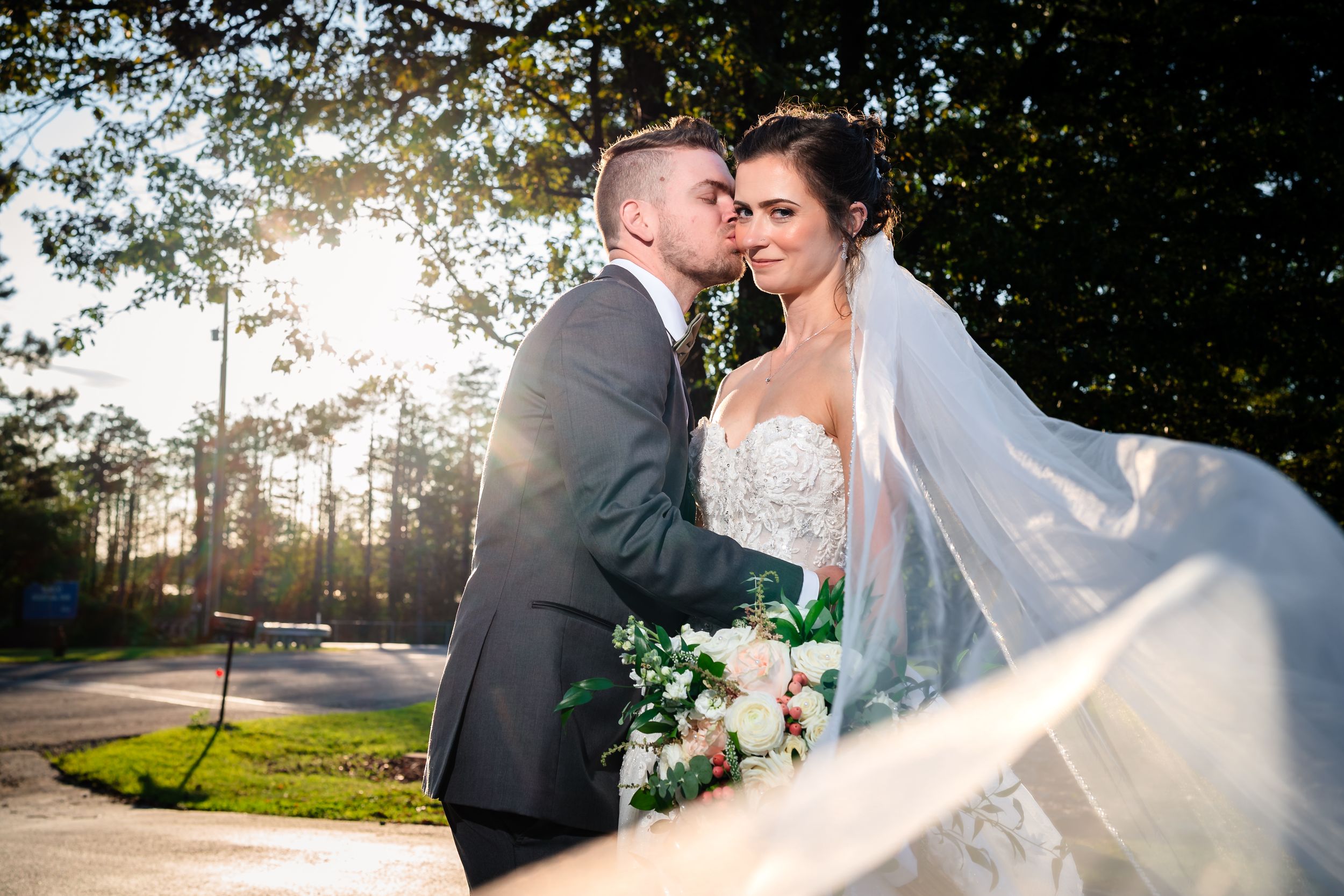 A romantic outdoor wedding portrait with sunlight filtering through trees.