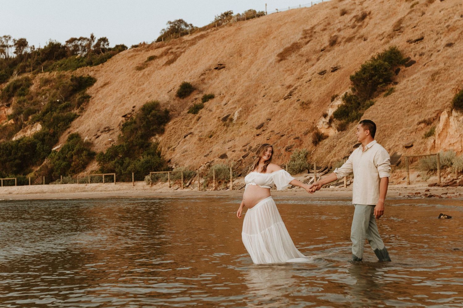 Couple wades in ocean water against cliff backdrop during golden hour.