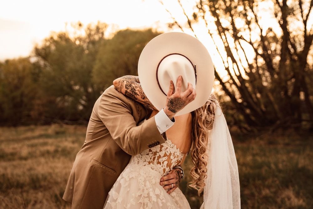 Bride and groom sharing a romantic kiss hidden behind a wide-brim hat at golden hour in a rustic outdoor setting.