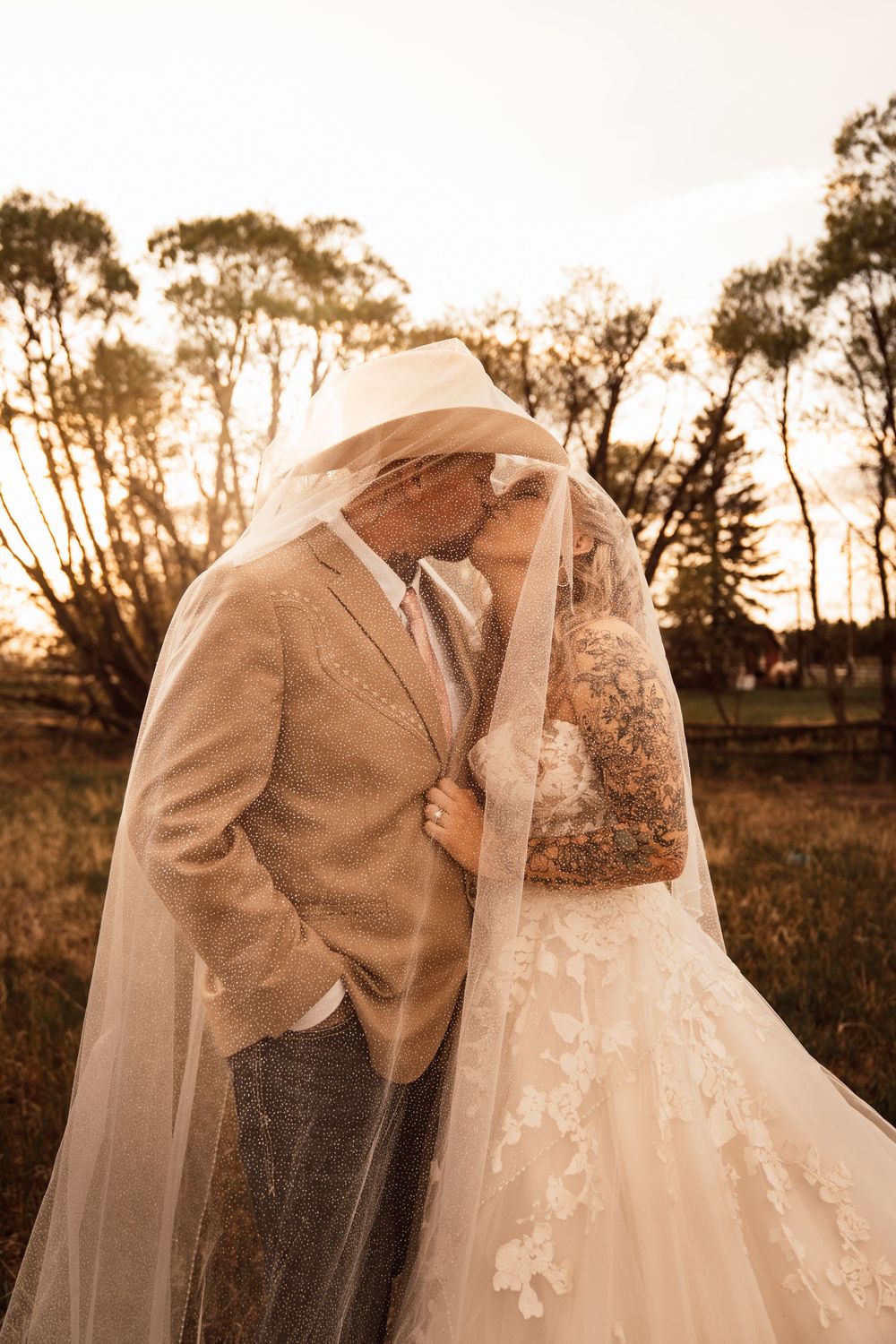 Bride and groom kissing under wedding veil at sunset with trees in background.