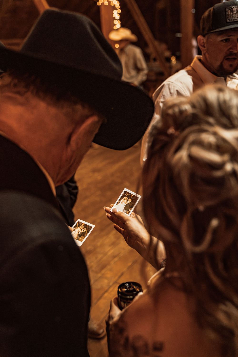 Wedding guests examine Polaroid photos at a rustic barn reception, with a man in a cowboy hat and bride with updo hairstyle.