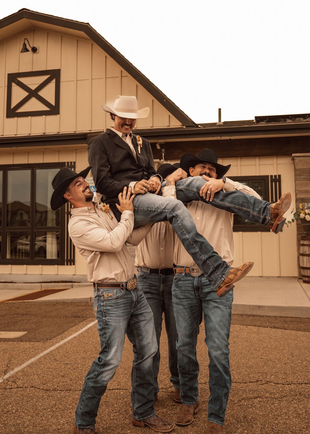 Cowboys in western attire lifting someone in a playful group pose outside a barn.