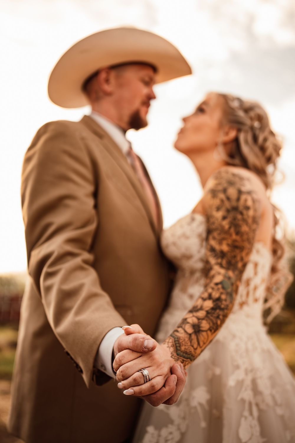 A bride with tattoos and groom in cowboy hat hold hands at golden hour wedding ceremony.