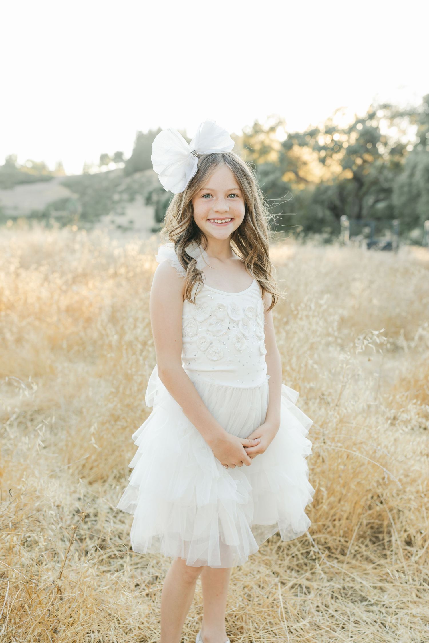 A young child wearing a white lace dress poses in a sunlit field of golden grass during a photo session captured by Chelsee Rawe Photograph.