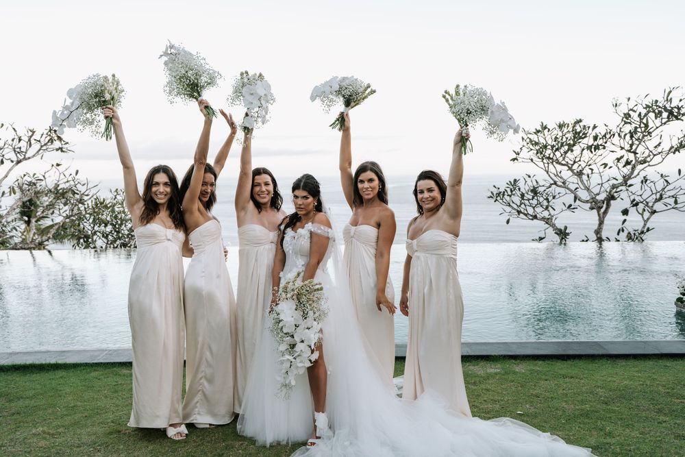 A wedding party in white dresses poses together near majestic trees at an outdoor venue overlooking a scenic view.