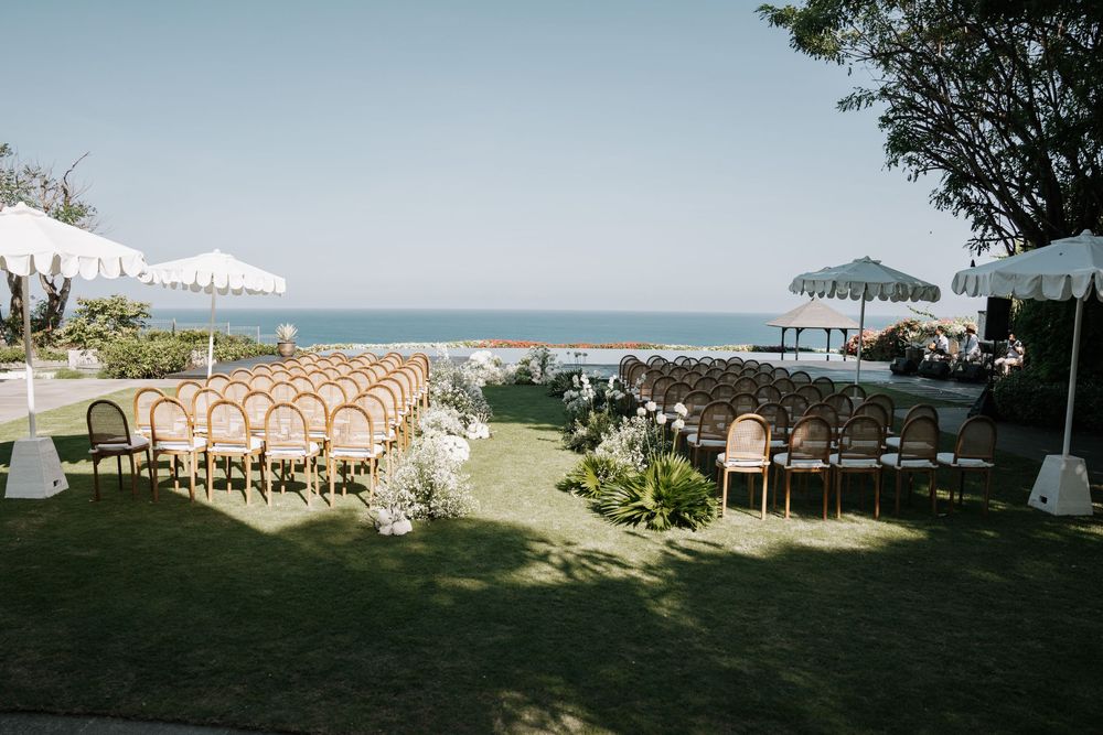 Outdoor wedding ceremony setup with white chairs and umbrellas on a grassy lawn overlooking the ocean.