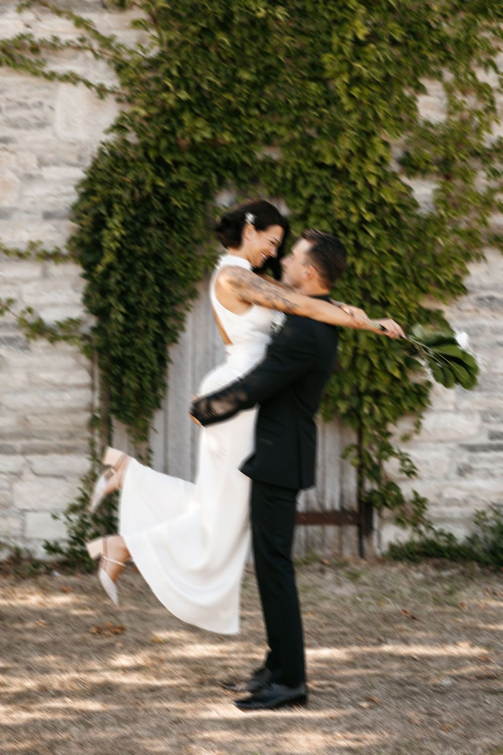 A couple in wedding attire embraces against an ivy-covered stone wall, with the bride's dress flowing in a romantic spin.