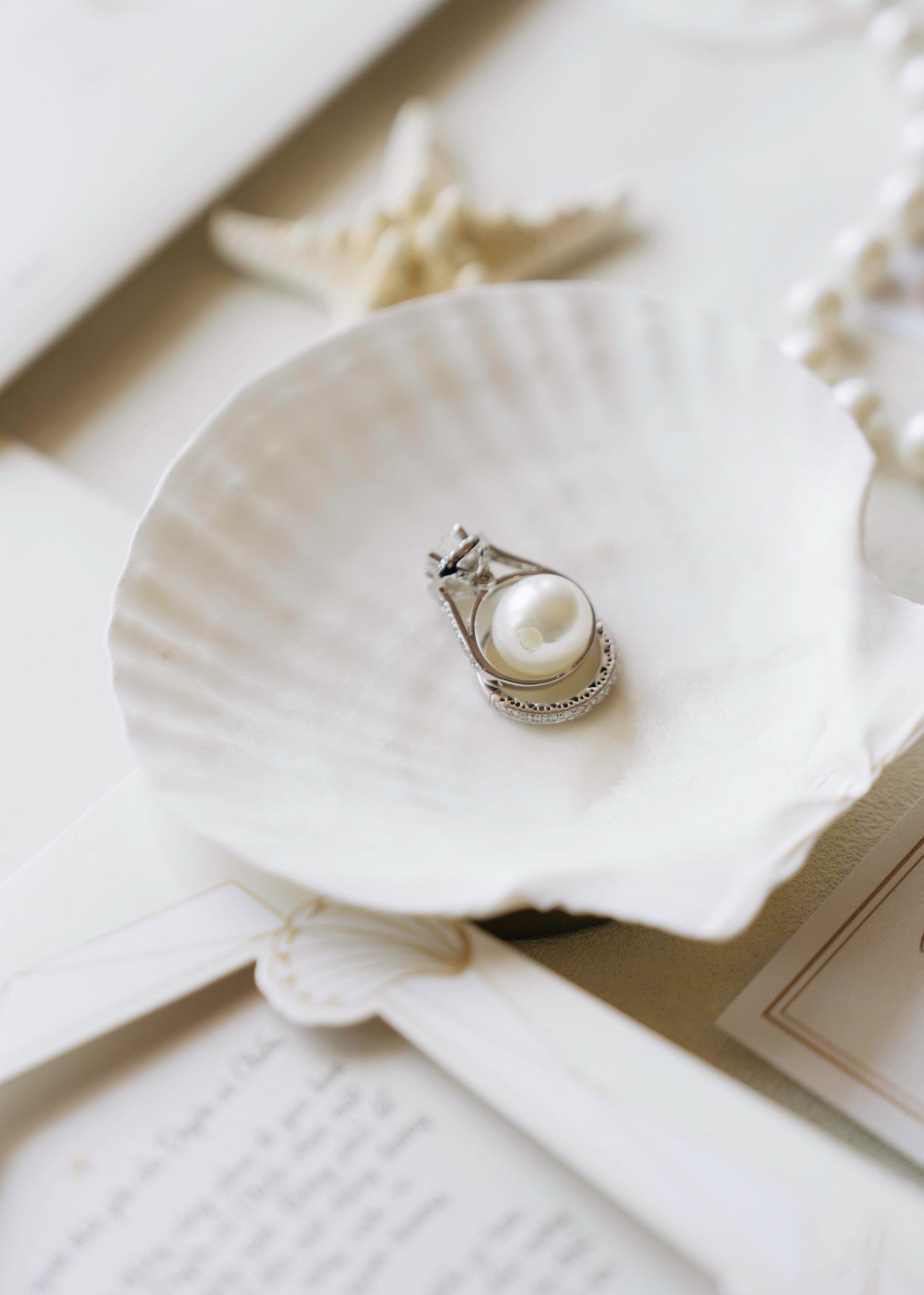 Pearl and diamond engagement ring displayed in a white scallop seashell on wedding stationery.