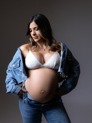 Pregnant woman in white lingerie and denim jacket poses against dark backdrop in maternity photoshoot series.