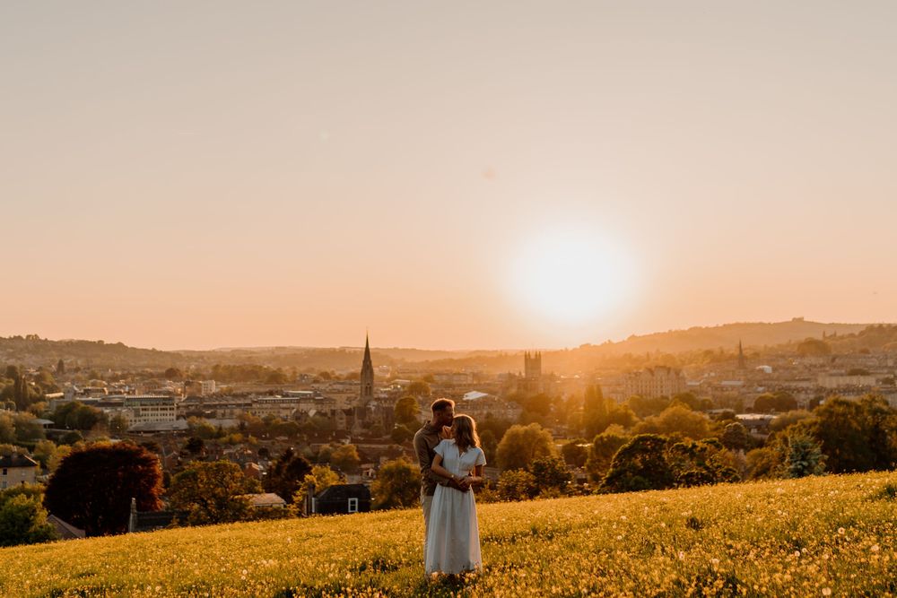 A couple embraces on a hilltop meadow overlooking a city at golden hour sunset with church spires visible below.