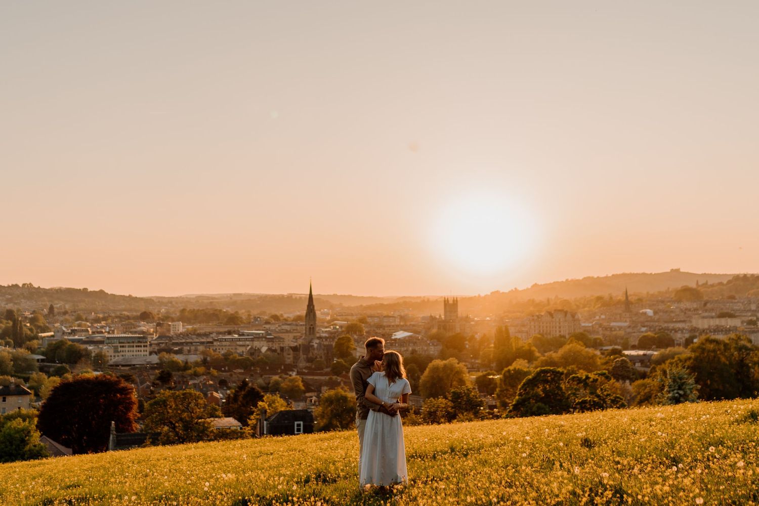 A couple embraces on a hilltop meadow overlooking a city at golden hour sunset with church spires visible below.