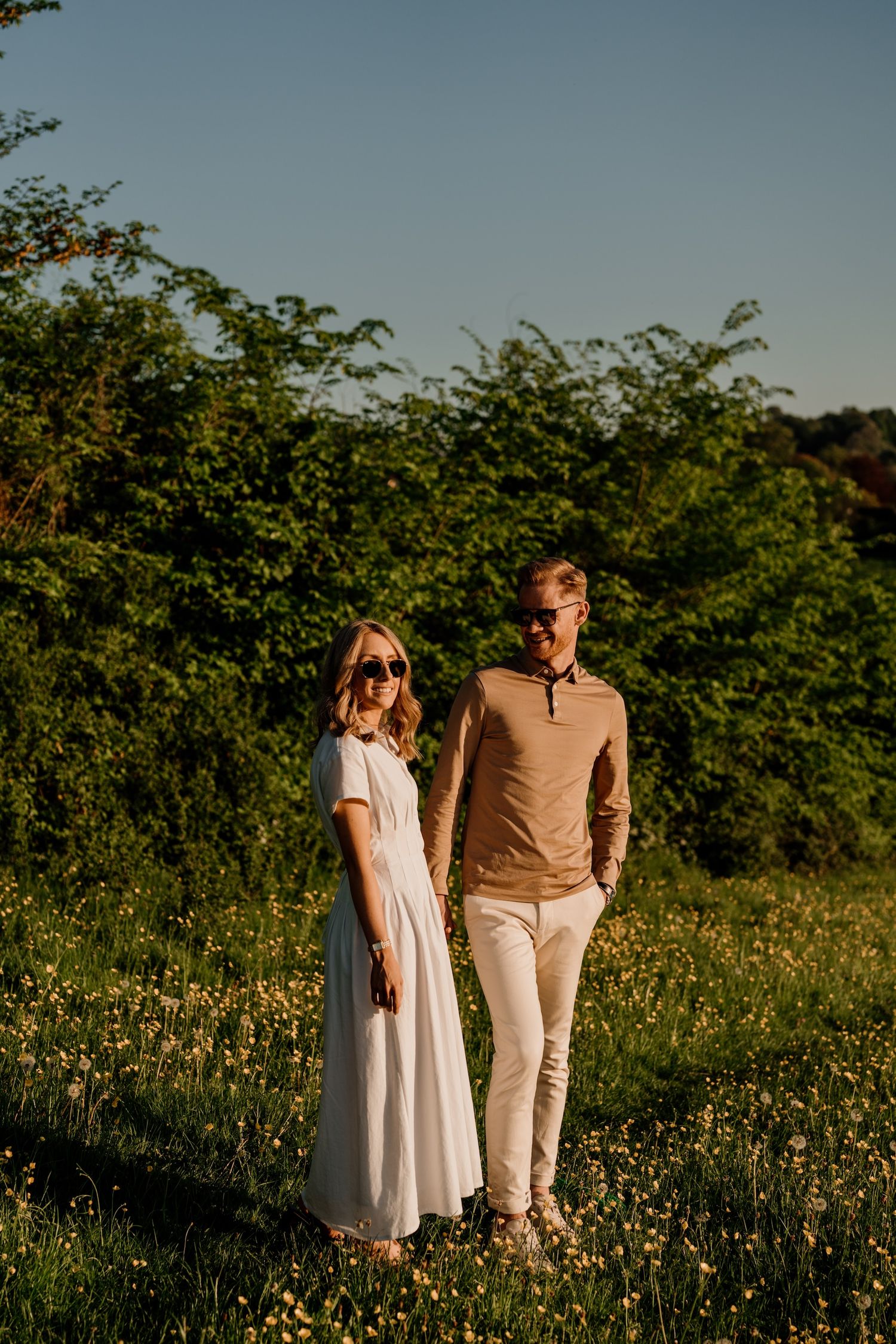 A couple walks together through a sunny meadow filled with wildflowers during golden hour.