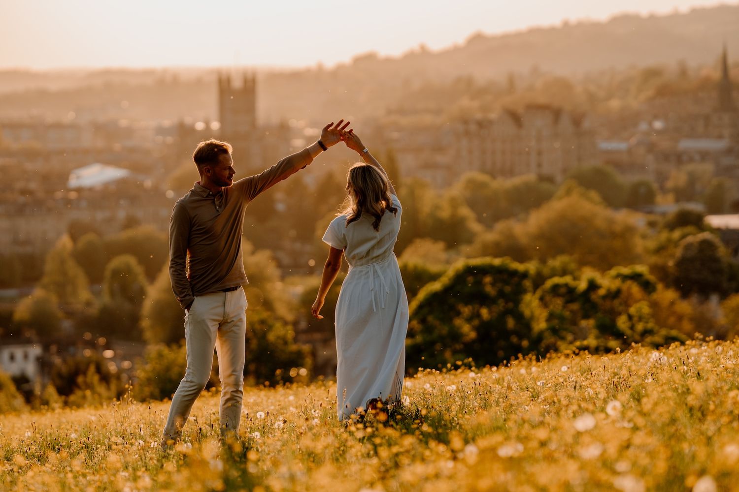 Two people running through a golden field during sunset with a cityscape visible in the background.