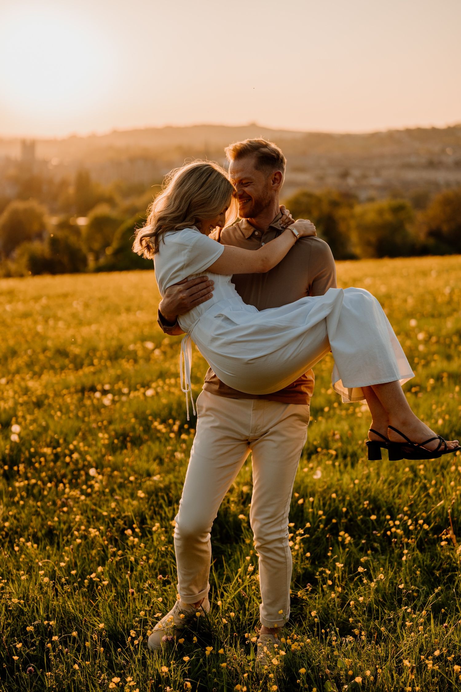 Couple in white outfits dancing playfully together in a sunlit meadow of wildflowers at golden hour.