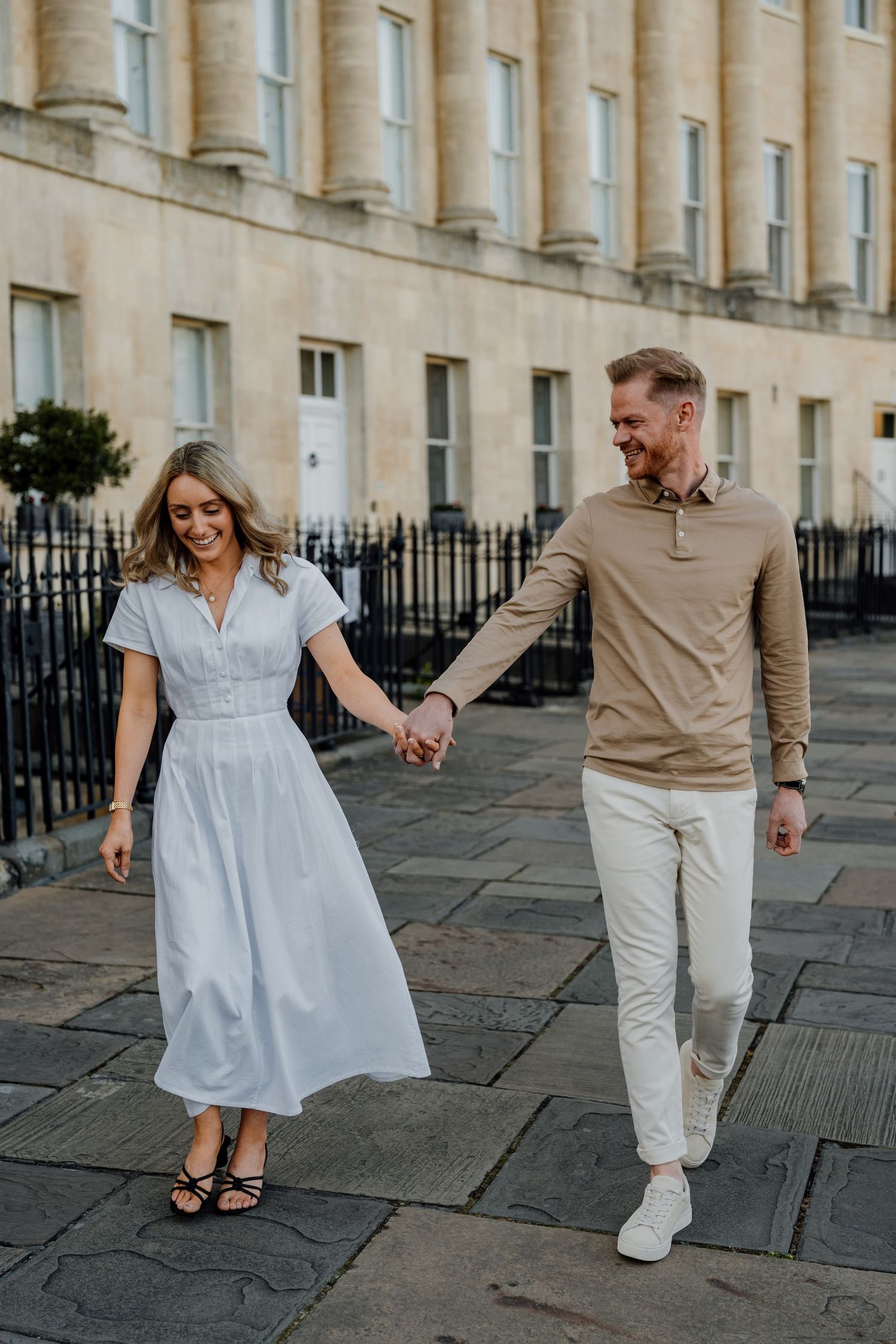 A couple in coordinated outfits walk hand in hand along a historic street with Georgian architecture in Bath, England.