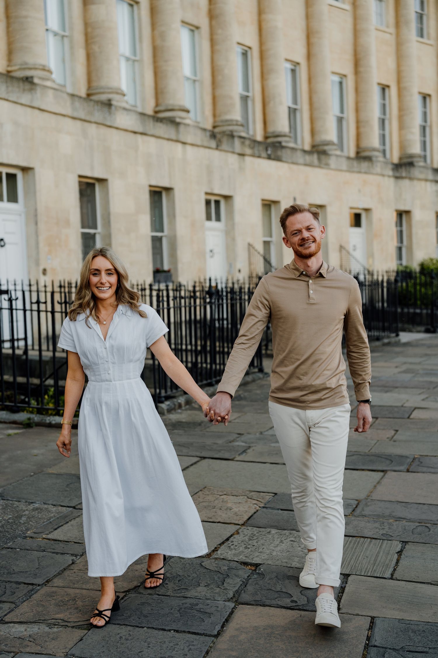 A couple in white and beige outfits holds hands while strolling along a historic stone-paved street.