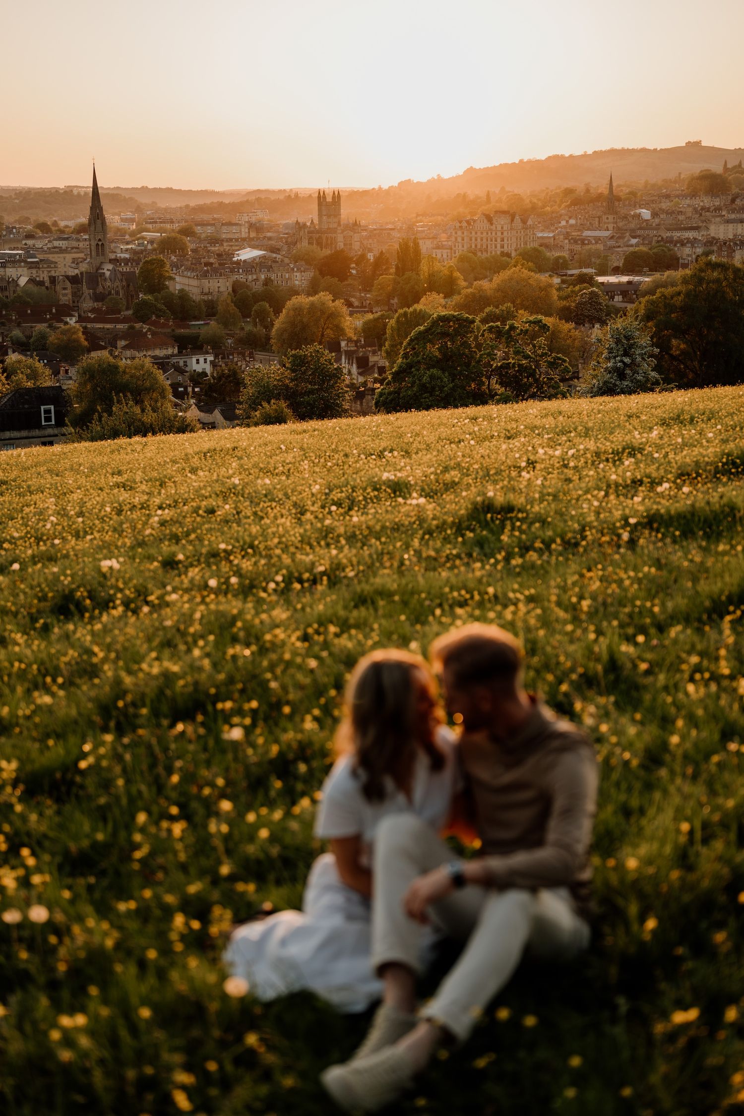 Couple sitting in a flower-filled meadow overlooking a historic city skyline with church spires at golden sunset.