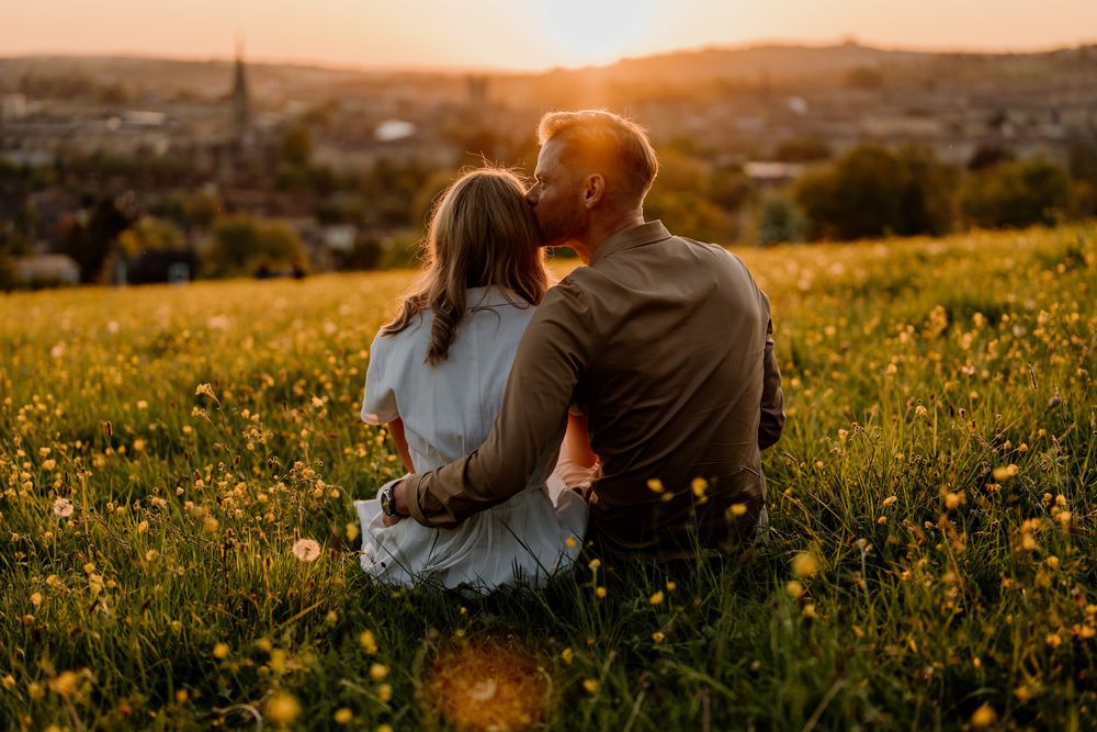 A couple sits in a wildflower meadow overlooking a town at golden hour as the sun sets behind hills in the distance.