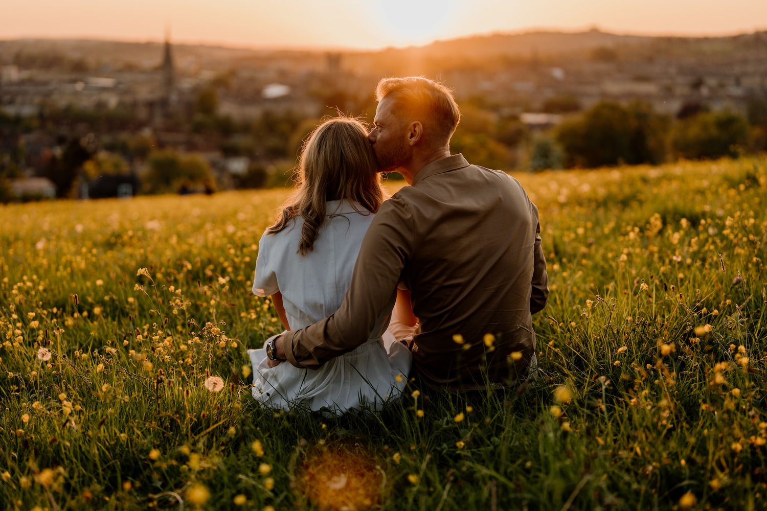 A couple sits in a wildflower meadow overlooking a town at golden hour as the sun sets behind hills in the distance.
