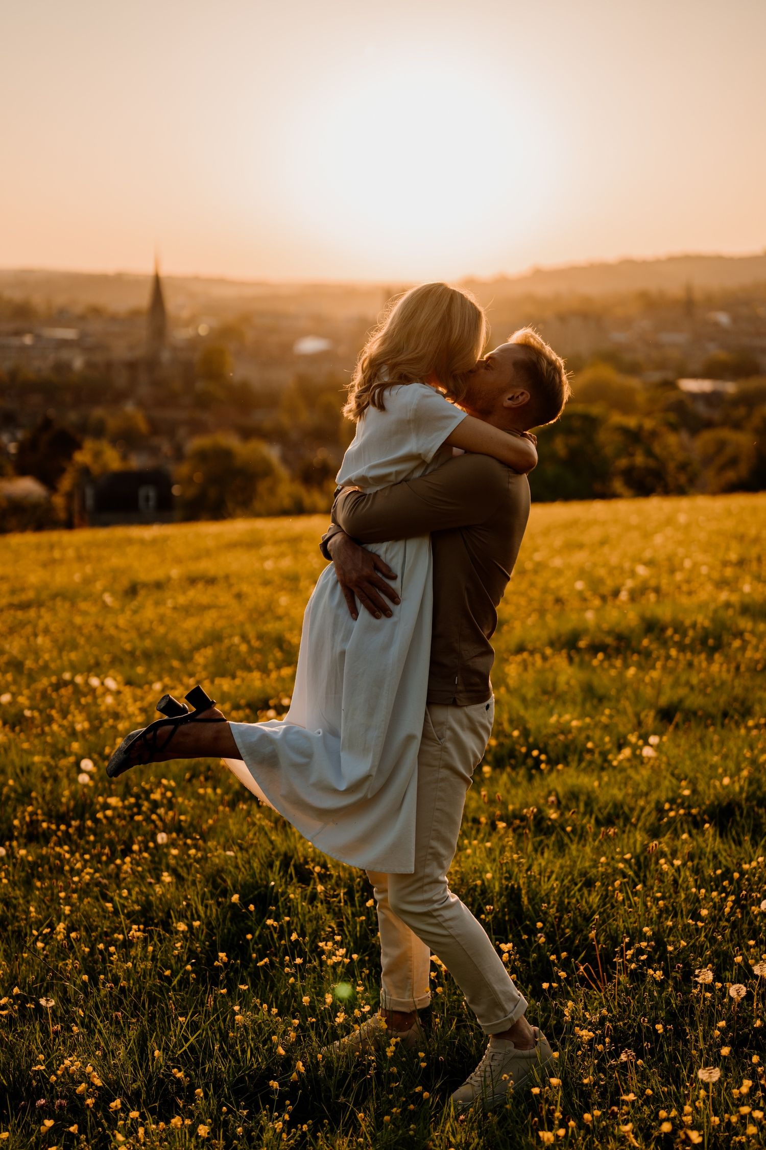 Two people embrace in a golden meadow with wildflowers at sunset as warm sunlight illuminates the pastoral landscape.