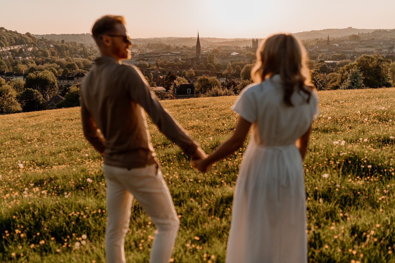 Silhouetted figures hold hands while walking through a meadow during sunset with cityscape in background.