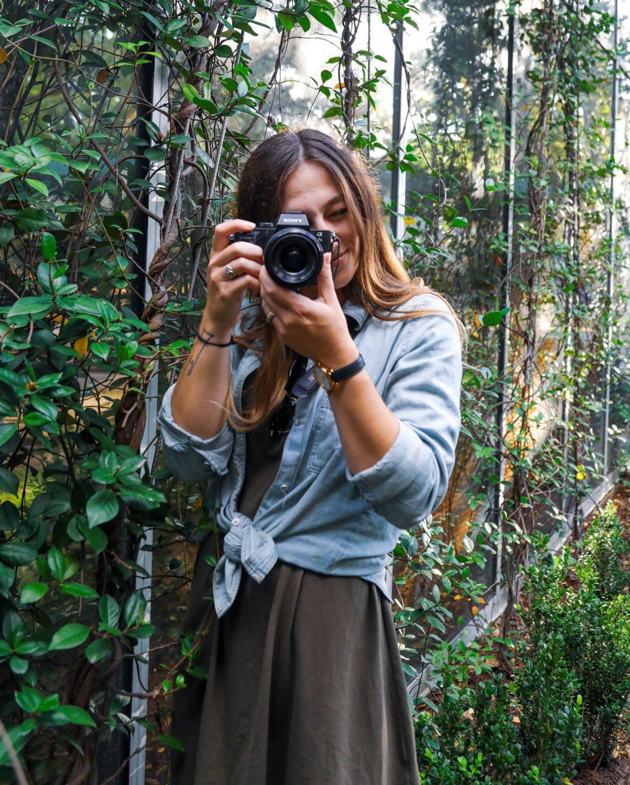 A photographer in a tied grey shirt and skirt takes pictures among green foliage.