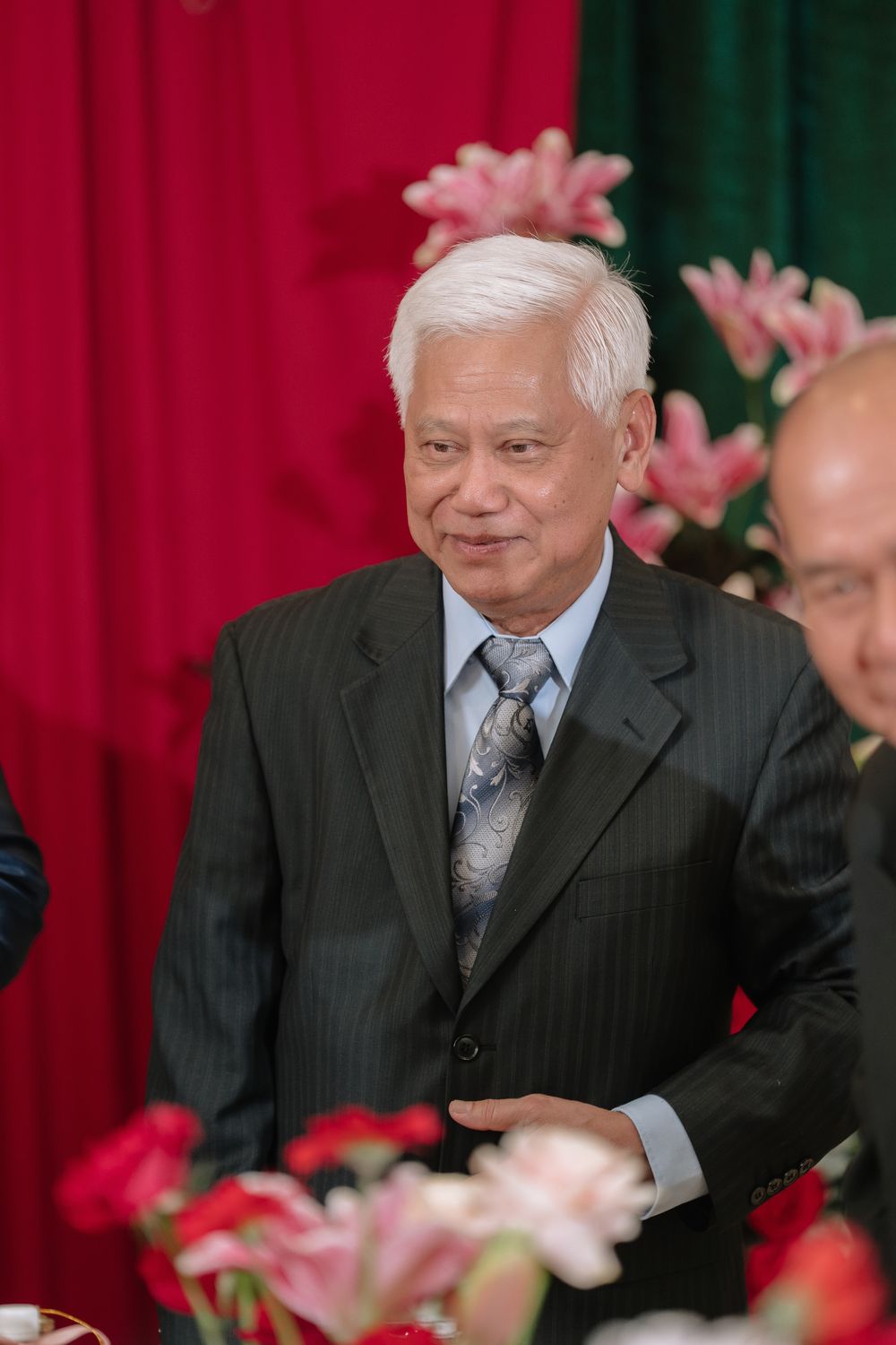 Elderly Asian man in dark suit and tie smiling at a formal event with floral decorations.