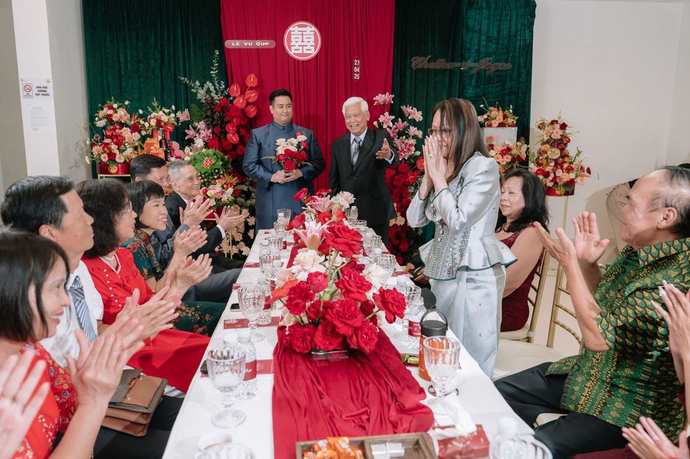 Guests applauding at a traditional Vietnamese wedding ceremony with red floral table decorations.