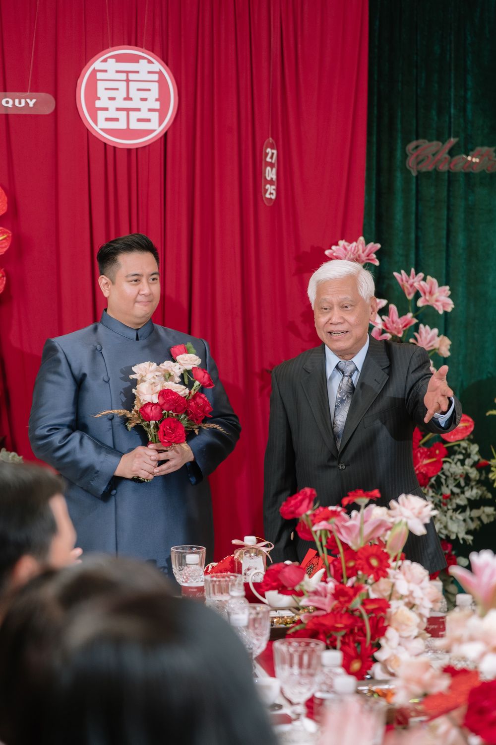 Two men standing at a Vietnamese wedding ceremony with red floral decorations and double happiness symbol.