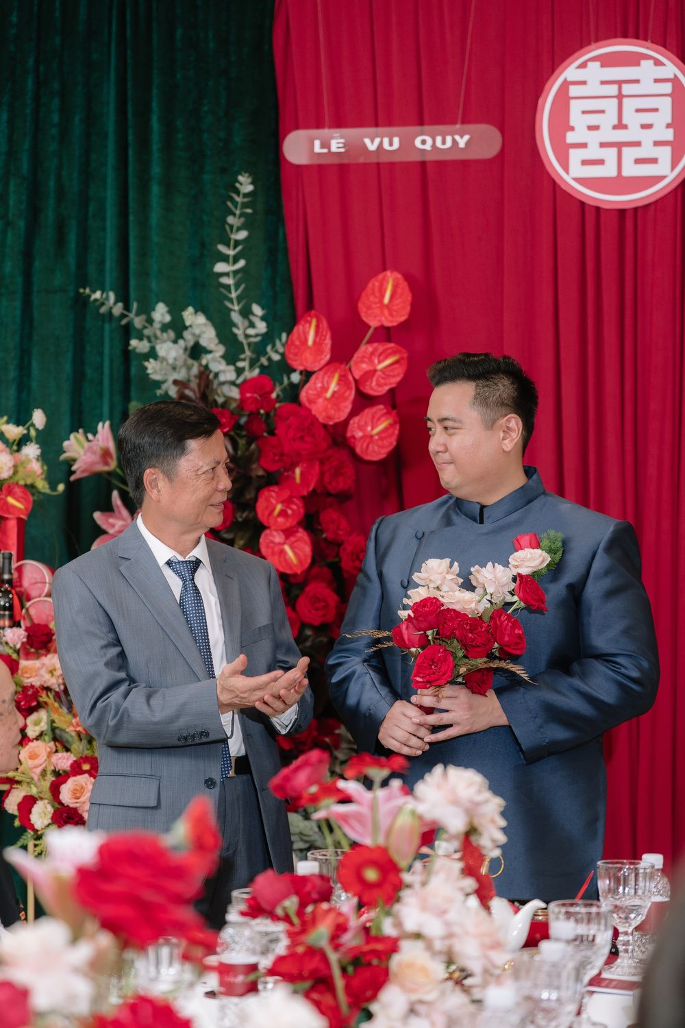 Two men conversing at a Vietnamese wedding ceremony with red anthurium flowers and happiness symbol backdrop.