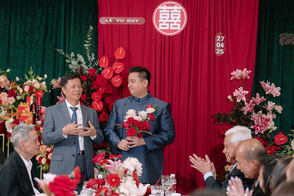 Two men speaking at a Vietnamese Le Vu Quy wedding ceremony with floral decorations and applauding guests.