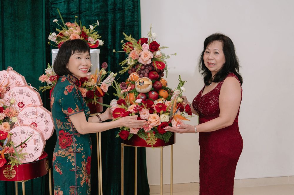 Two women in elegant qipaos exchange a vibrant floral arrangement at a traditional Chinese celebration.