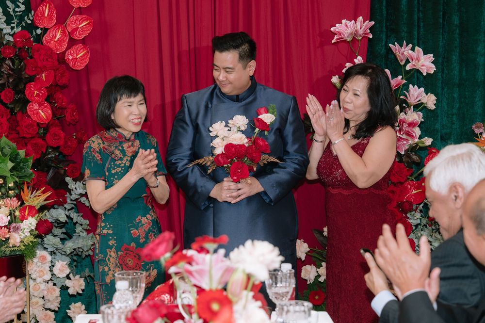 A groom stands between two women in traditional Chinese wedding attire amid red floral decorations.