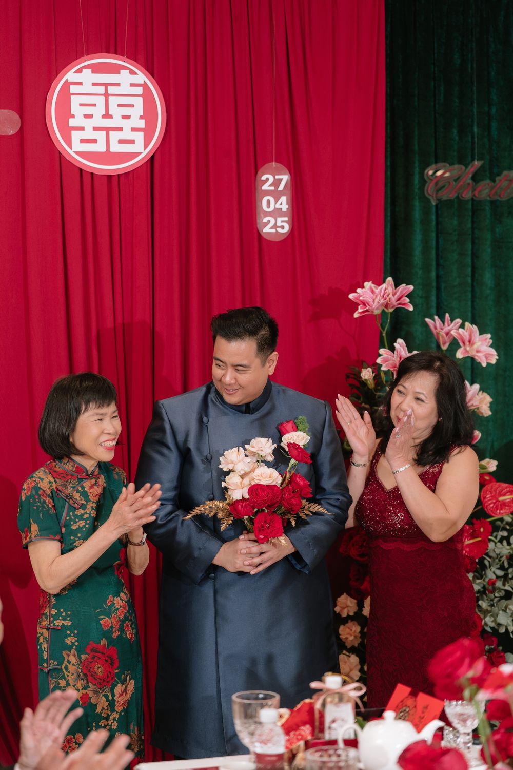 A groom in blue tang suit holds bouquet flanked by two women at a Chinese wedding ceremony.