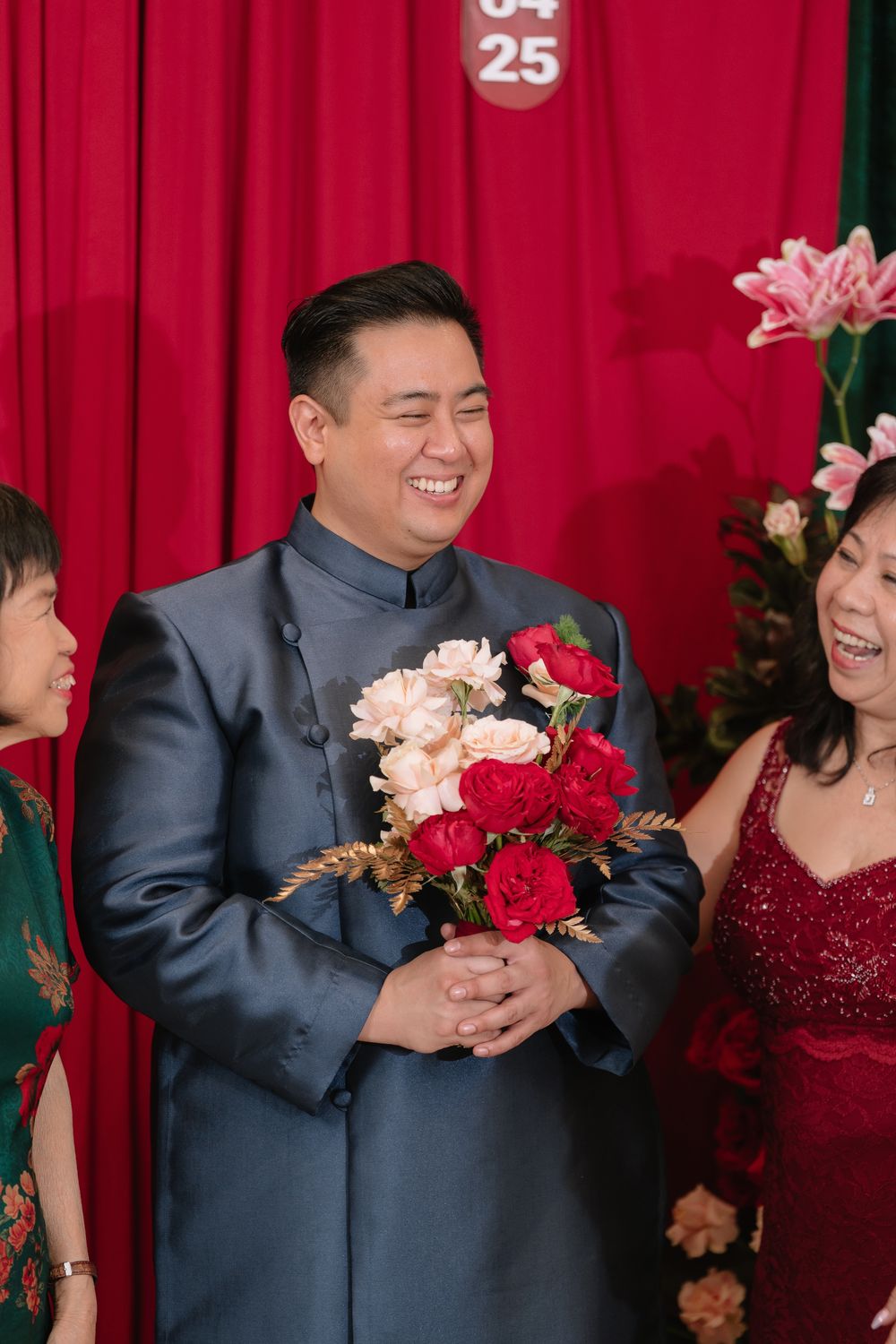 A smiling groom in a blue tang suit holds red and pink roses at a Chinese wedding celebration.