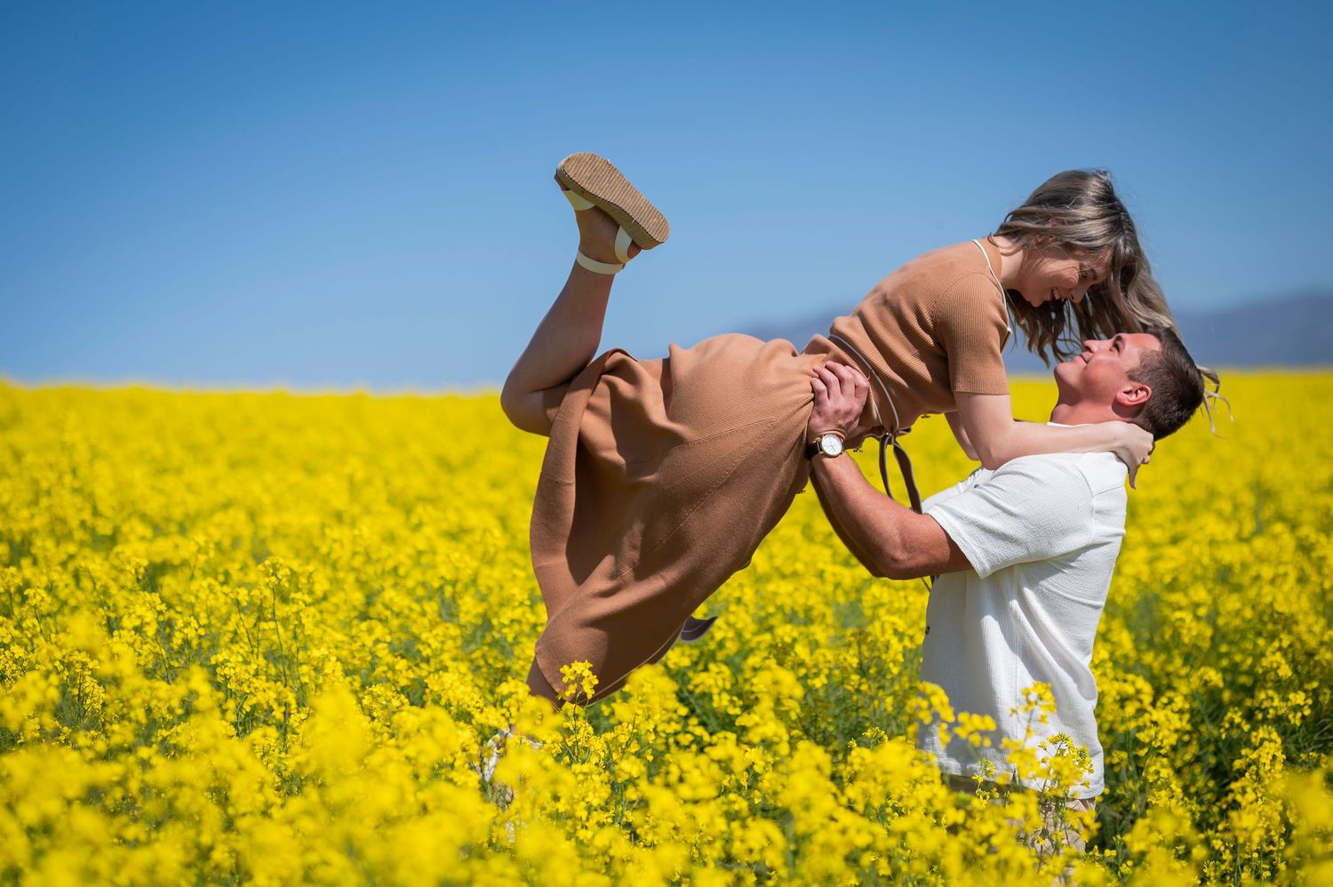 Two people share a romantic moment while standing in a vibrant yellow canola field against a bright blue sky.