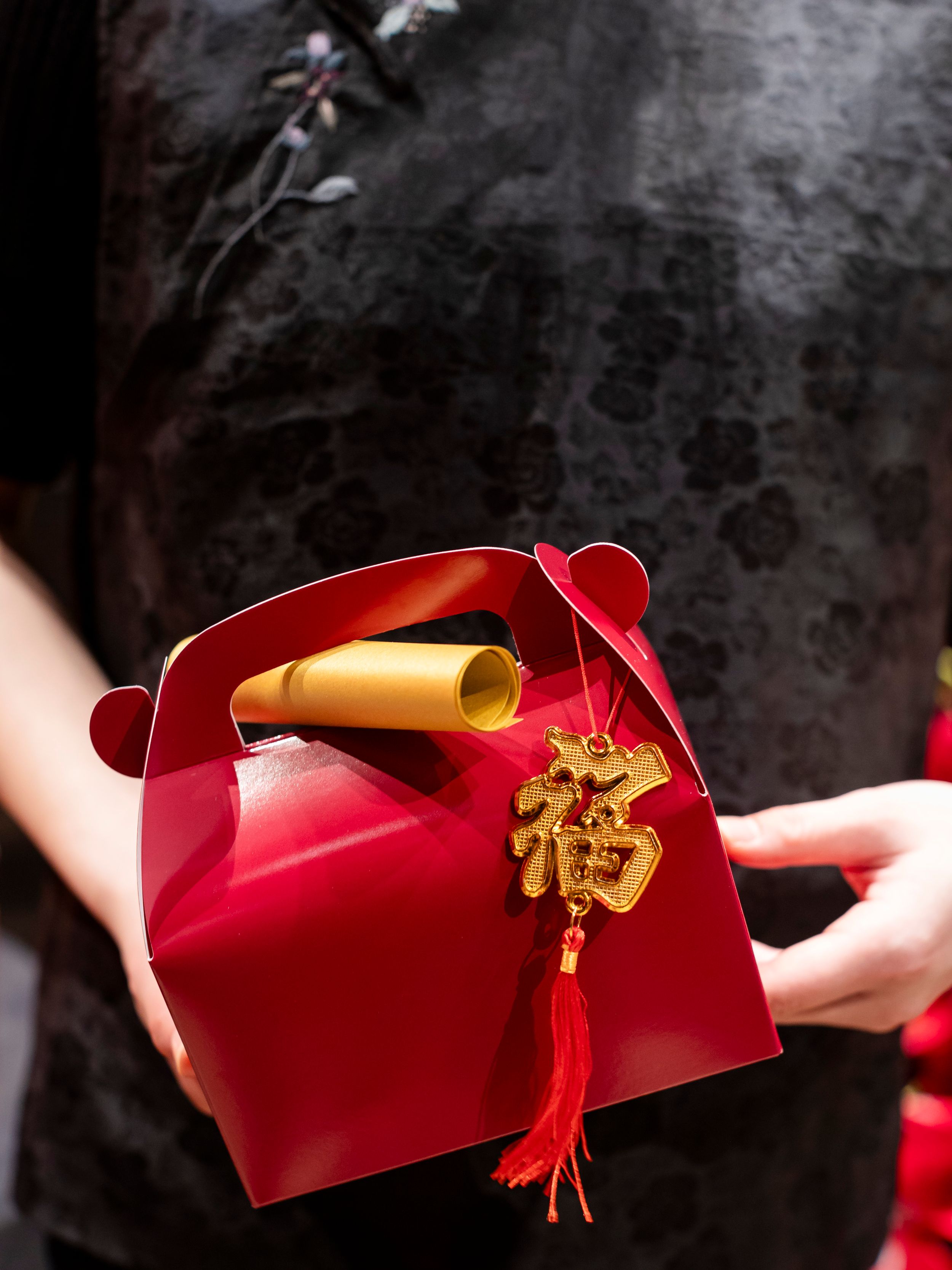 Red gift box with gold scroll and tassel ornament held against dark background.
