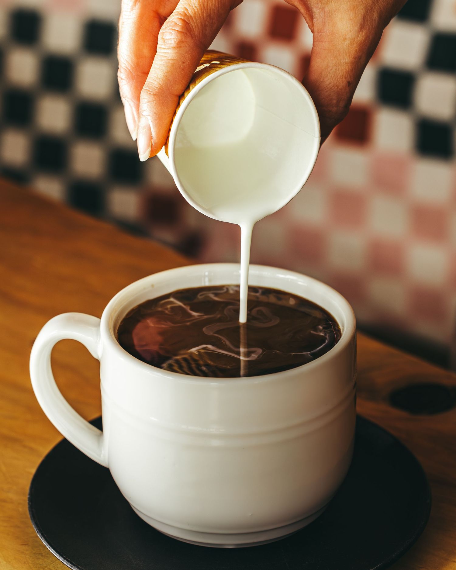 White ceramic cup being filled with milk pouring into dark coffee on wooden surface with checkered background.