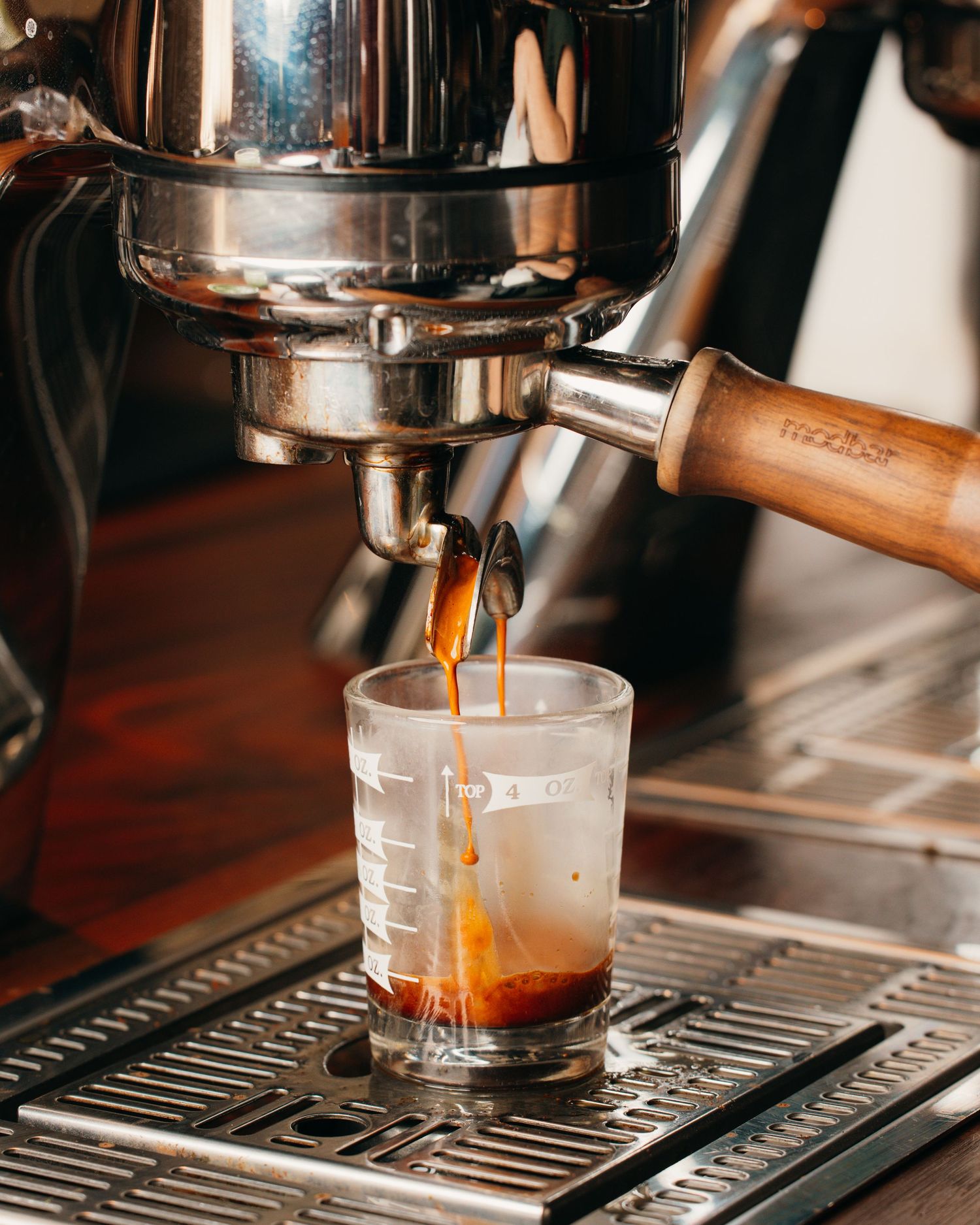 Espresso machine brewing fresh coffee into a clear glass measuring cup on metal grate surface.