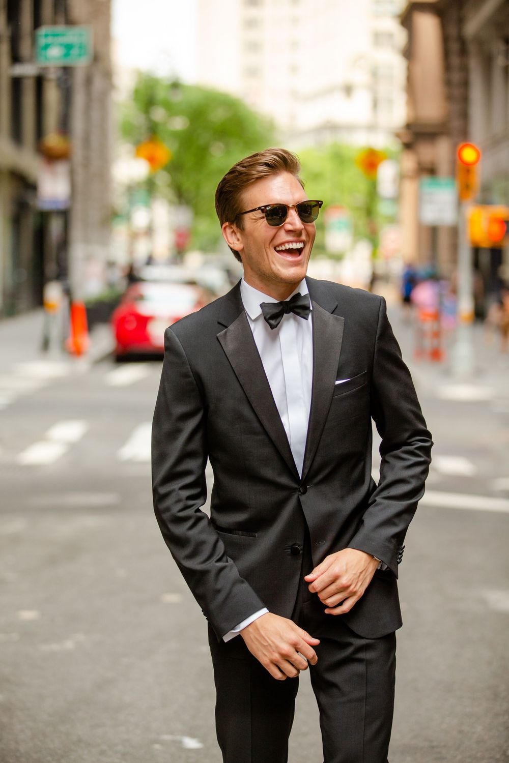 Stylish groom in black tuxedo and bow tie smiles while standing on city street.