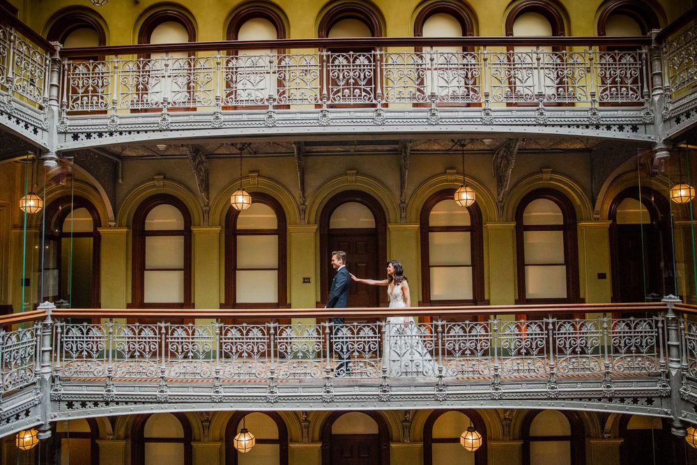 An elegant wedding couple stands on an ornate Victorian balcony with arched windows and decorative railings.